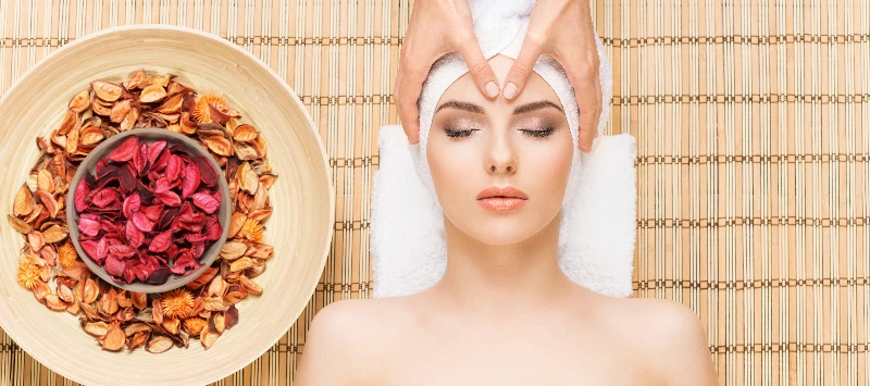 A imagem apresenta, em plano médio e visão lateral, uma mulher deitada de bruços sobre uma maca de massagem branca com estrutura e acabamento em madeira clara. Ela está recebendo uma massagem nas costas de uma profissional em um ambiente de spa.
