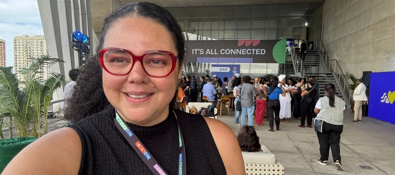 "Pessoa usando óculos vermelhos e blusa preta sem mangas tira uma selfie em um evento social. Ela usa crachá com várias etiquetas. Ao fundo, há um grupo de pessoas reunidas perto de mesas e escadas sob uma estrutura moderna de concreto. Um banner grande c