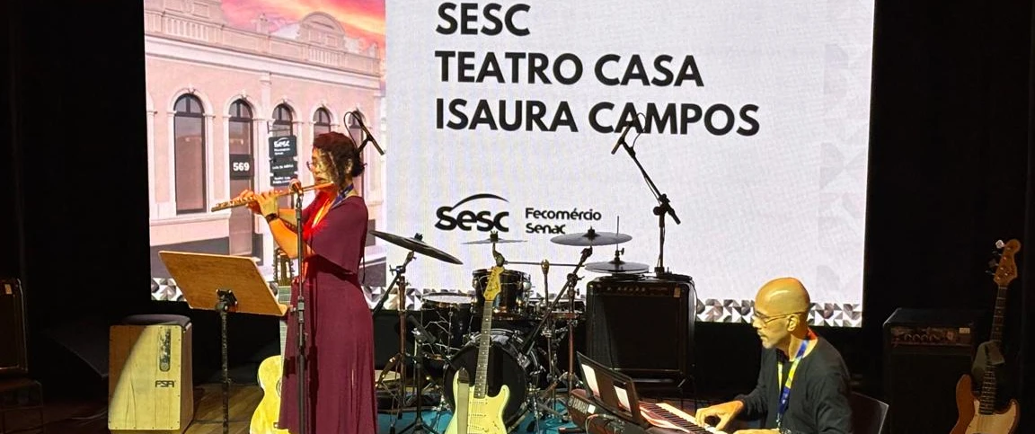 Fotografia de um palco de teatro com iluminação em tons quentes. No centro-esquerda, uma pessoa está em pé tocando flauta diante de um pedestal com partitura e um microfone. À direita, outra pessoa está sentada tocando teclado eletrônico. 
