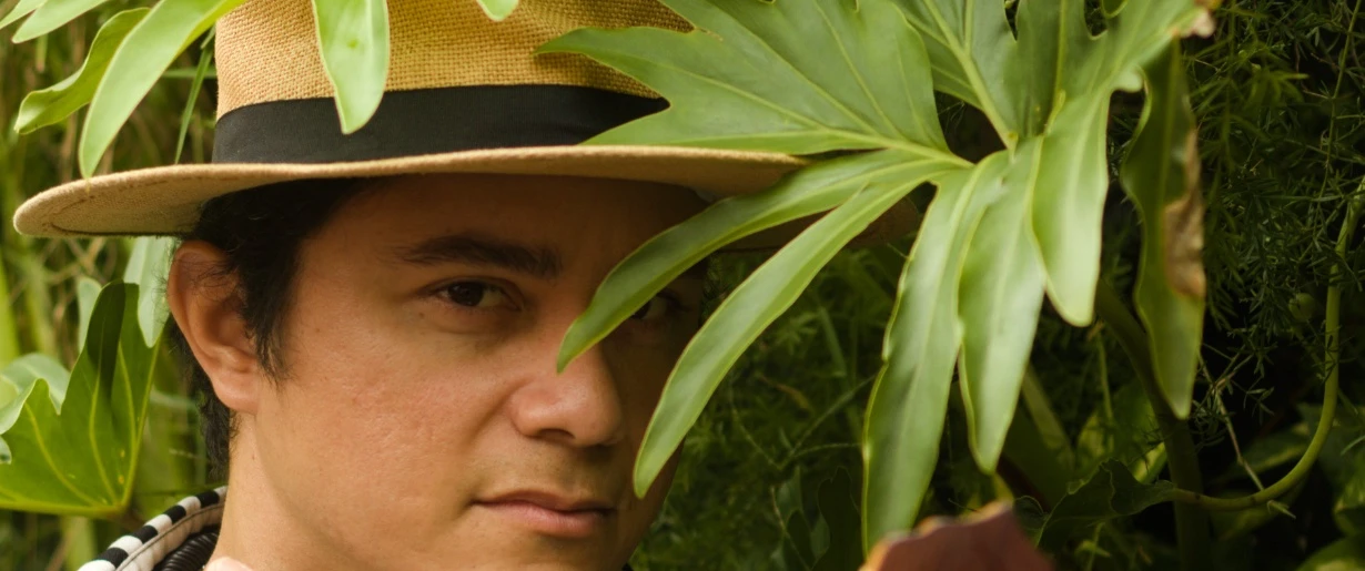 Pessoa com chapéu de palha entre folhagens tropicais, vestindo camisa listrada em preto e branco, com um violão em primeiro plano. A imagem destaca a integração entre a vegetação verde e os elementos musicais.