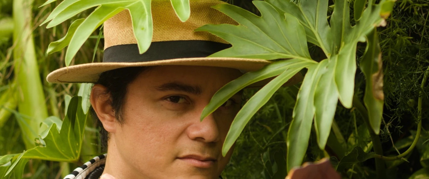 Pessoa com chapéu de palha entre folhagens tropicais, vestindo camisa listrada em preto e branco, com um violão em primeiro plano. A imagem destaca a integração entre a vegetação verde e os elementos musicais.