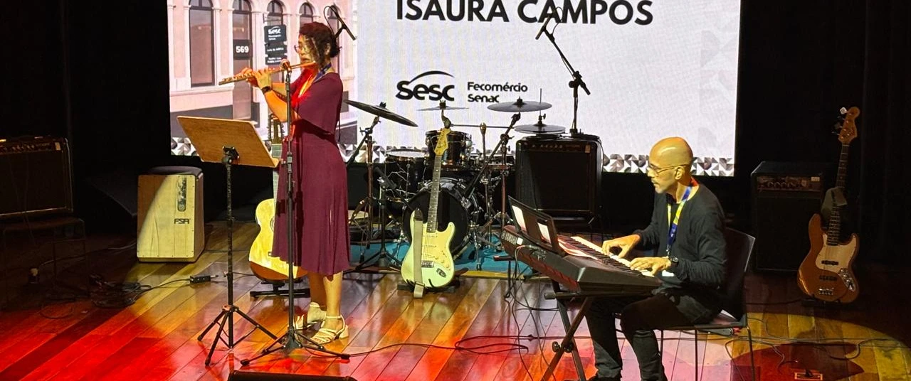 Fotografia de um palco de teatro com iluminação em tons quentes. No centro-esquerda, uma pessoa está em pé tocando flauta diante de um pedestal com partitura e um microfone. À direita, outra pessoa está sentada tocando teclado eletrônico. 