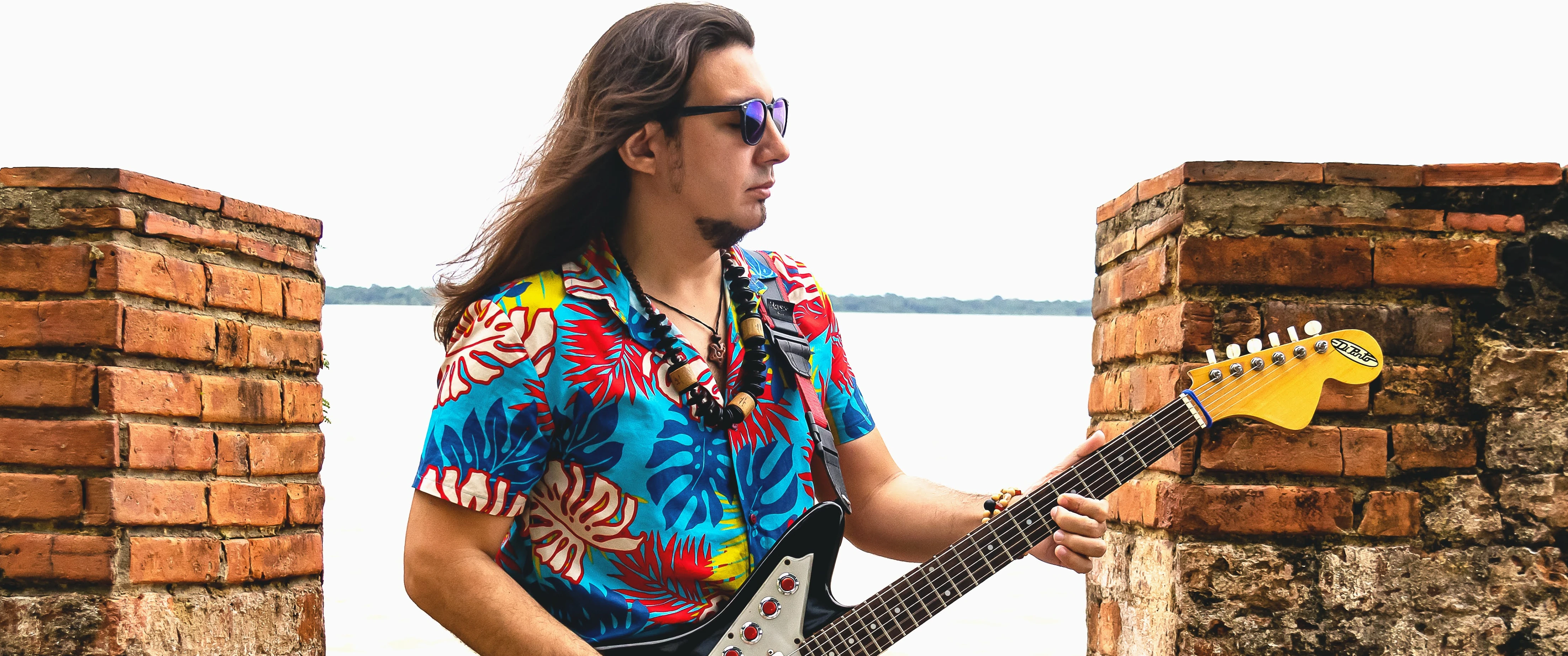 O músico está com camisa colorida, óculos escuros e guitarra na mão.