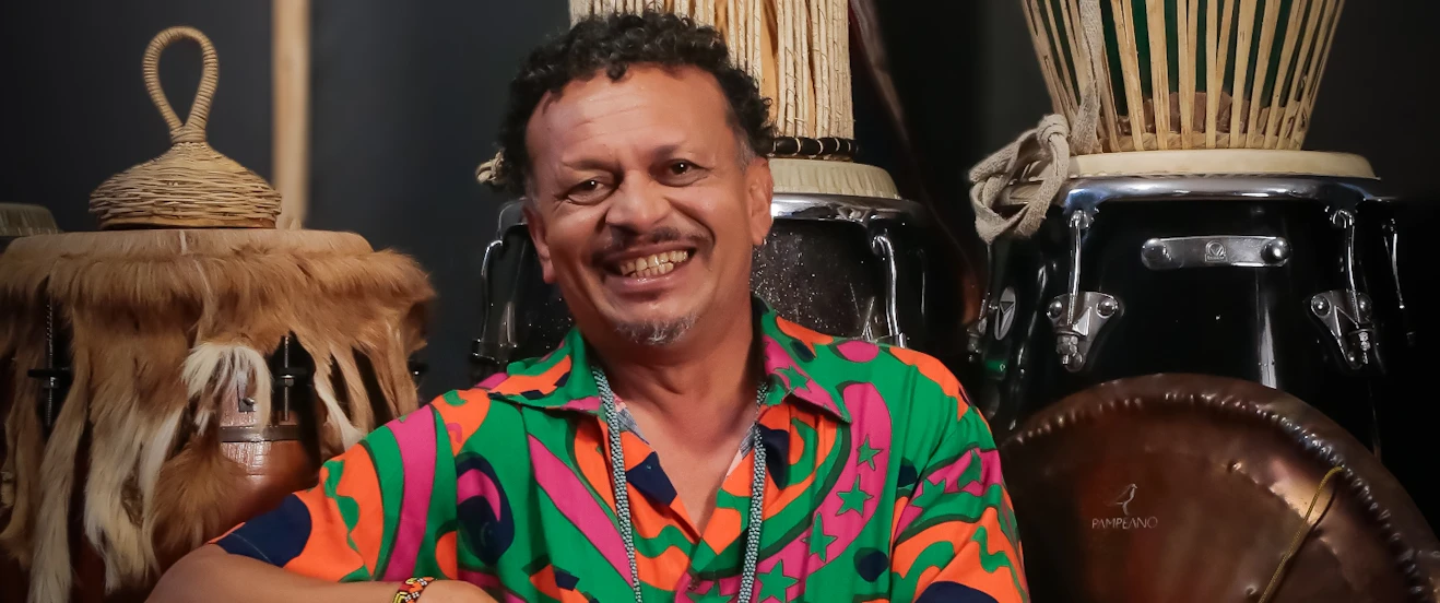 Descrição para o site do Sesc  Pessoa sentada entre diversos instrumentos de percussão, usando camisa colorida estampada. Em primeiro plano, um instrumento metálico, e ao fundo, tambores e outros instrumentos percussivos.