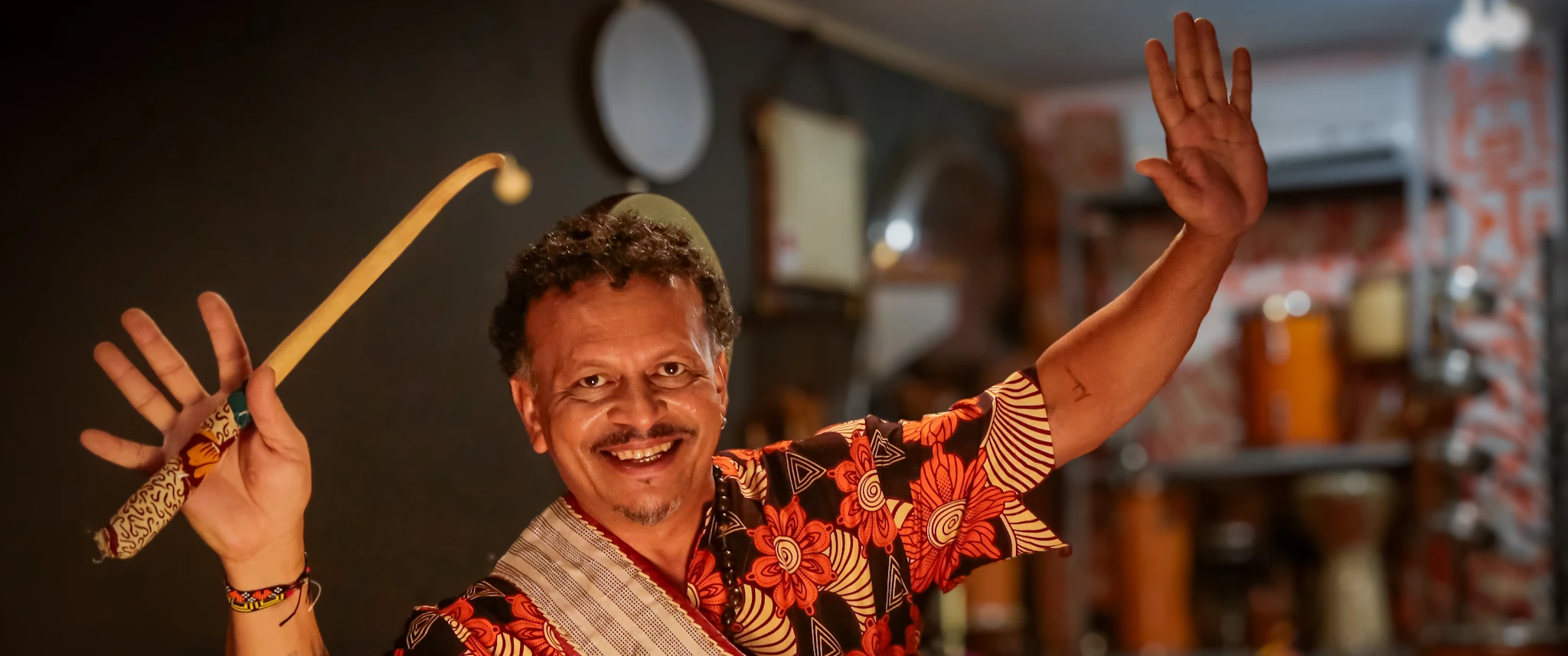 Descrição para o site do Sesc  Pessoa em destaque, segurando uma baqueta e um instrumento de percussão, usando camisa estampada em tons de vermelho, laranja, preto e bege, em um ambiente com diversos instrumentos musicais ao fundo.