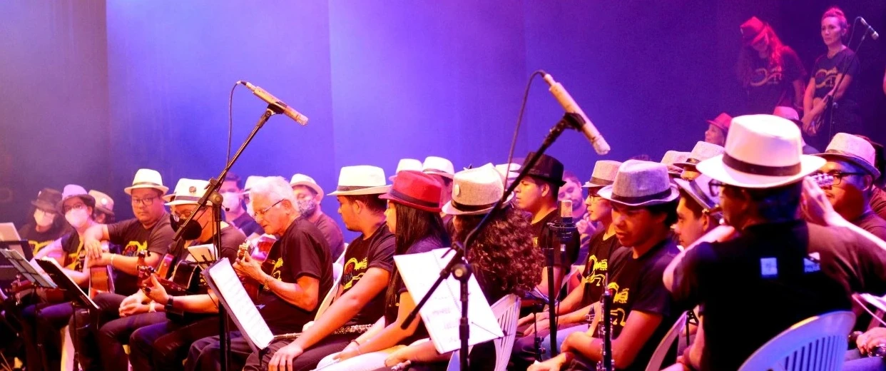 Grupo musical reunido em palco de teatro durante apresentação coletiva, com instrumentos, partituras e iluminação cênica em tons de azul e roxo. A imagem destaca a integração dos participantes e a valorização da prática musical em conjunto.